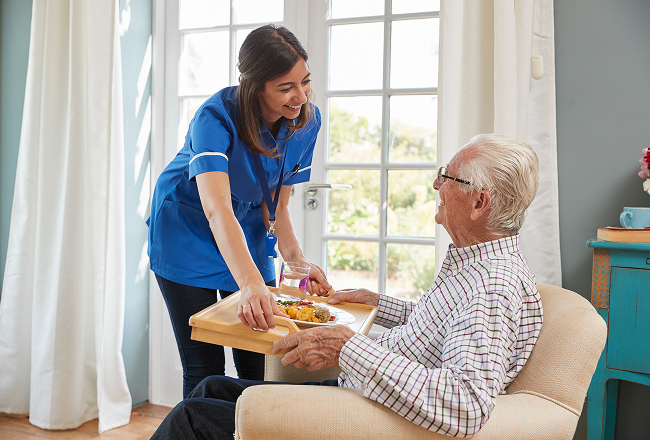 A caregiver in a blue uniform smiles warmly, serving a meal to an elderly man sitting in an armchair by a bright window, conveying care and kindness.