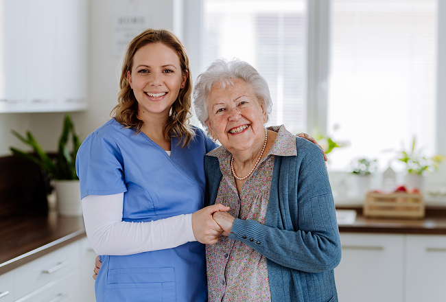 A smiling nurse in blue scrubs holds hands with a cheerful elderly woman in a cozy living room, conveying warmth and care.