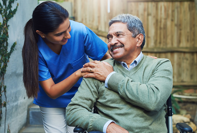 A nurse in blue scrubs gently holds an elderly man’s shoulder, who is smiling in a wheelchair.